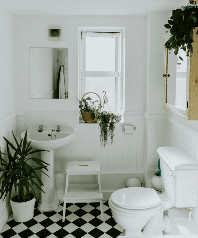 Bright white bathroom filled with lush greenery, featuring potted plants on the black & white checkered floor, hanging vines and a basket of potted plants on the window sill, and small hanging plant over the natural wood medicine cabinet, creating a fresh, natural ambiance.