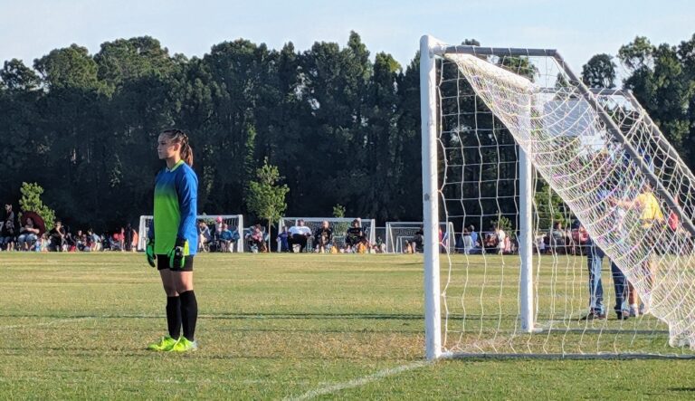 A soccer goalkeeper stands alert in front of the goal on a grassy field. The goalkeeper is wearing a blue and green jersey, black shorts, black knee-high socks, and bright green cleats. The net of the goal is visible on the right side of the image. In the background, a crowd of spectators is seated and watching the game, with a line of trees providing a natural backdrop.