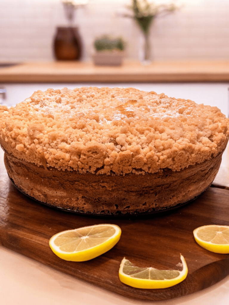 A golden-brown crumb cake with a crumbly topping is displayed on a wooden cutting board in a cozy kitchen setting. The cake is garnished with a light dusting of powdered sugar, and three lemon slices are arranged around it on the cutting board. The background shows a blurred view of kitchen countertops and decor.