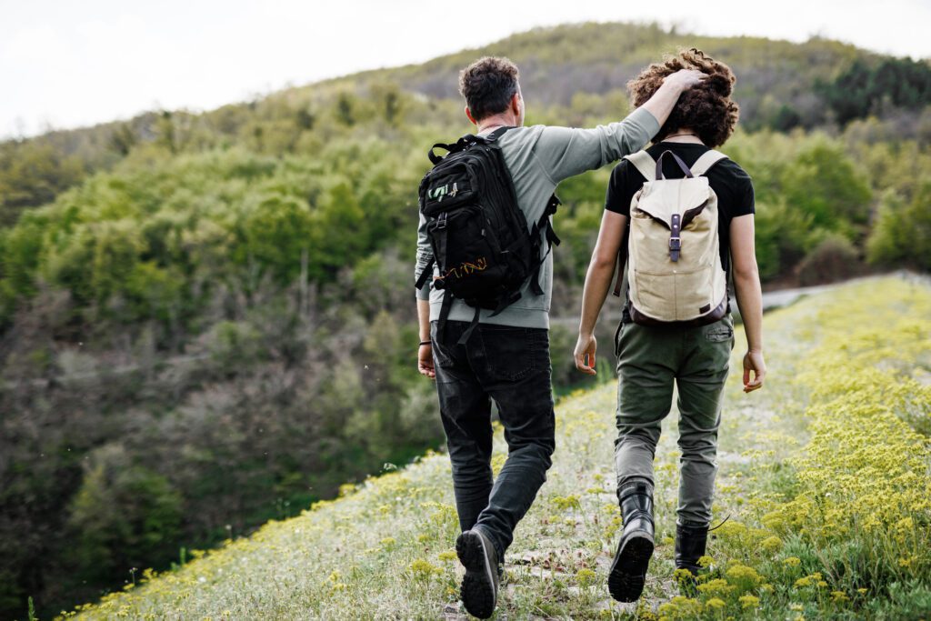 Two individuals, one older and one younger, walk along a scenic nature trail surrounded by greenery, symbolizing guidance and support. Both wear backpacks, and the older person affectionately places a hand on the younger one's head. This peaceful moment represents the journey of navigating the middle school years, highlighting the importance of mentorship, connection, and emotional support during a time of growth and self-discovery.